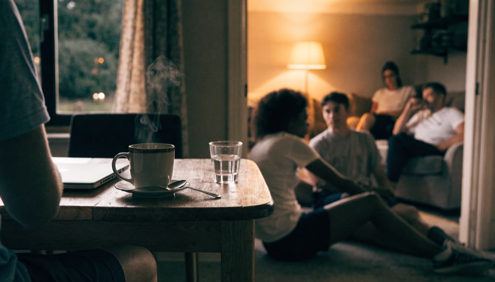 A volunteer's kitchen table at dusk — laptop closed, tea steaming, the family together in the warm room beyond