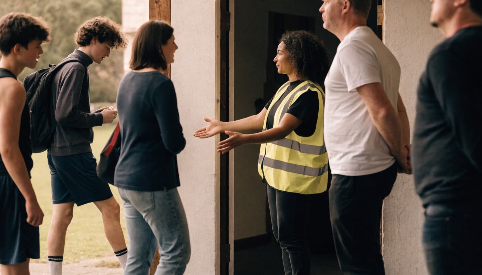 A welfare officer in hi-vis welcomes arriving players and parents at the clubhouse door, everything open and visible